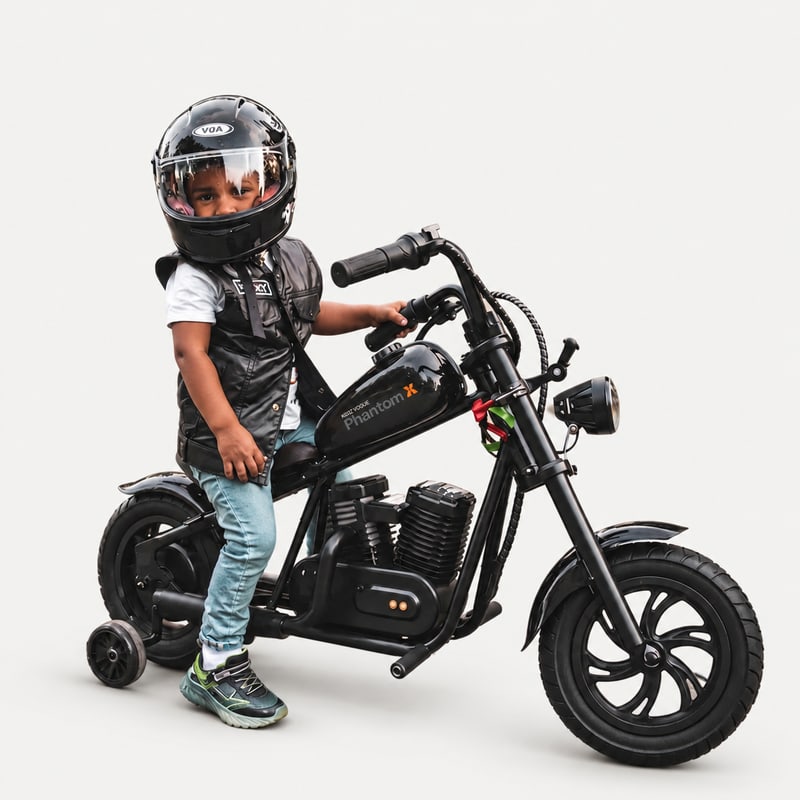 Black Phantom X electric ride-on bike with a helmeted child seated on it, shown from a front three-quarter angle on a light-grey background.
